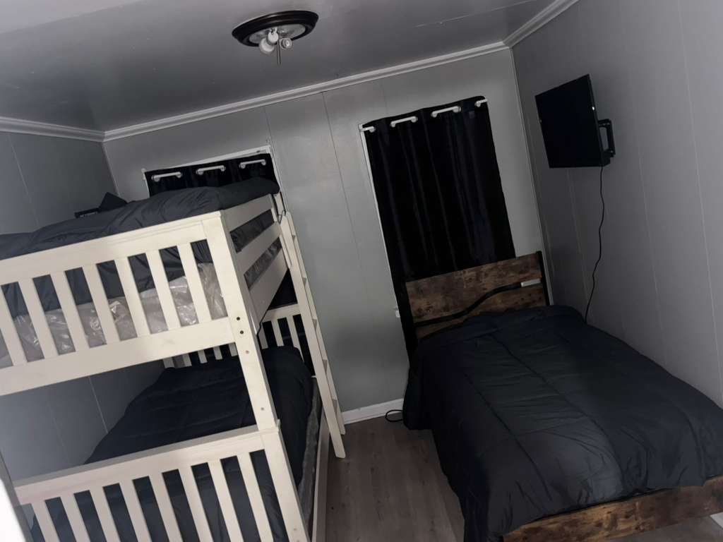 Compact bedroom featuring a white bunk bed, single bed, black curtains, and wall-mounted television.